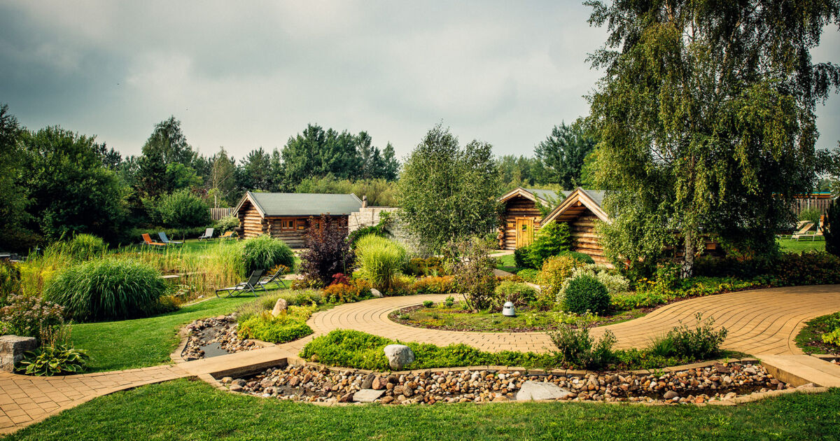 Im Bild wird unser großer idyllischer Saunagarten mit Bachlauf, Rundweg und Gartenteich gezeigt. Um den Gartenteich herum stehen Entspannungsliegen den Besuchern zur Verfügung. Große Bäume bieten Schatten und kleine Sträucher Sichtschutz.